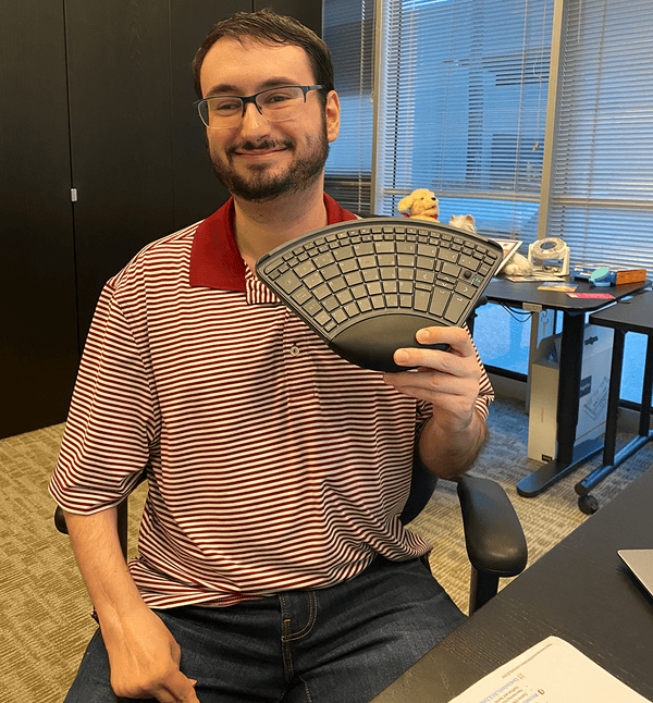 A man wearing glasses and a red striped shirt sits in an office, smiling and holding a split ergonomic keyboard.