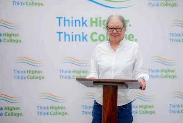 Debra Hart smiling at a podium in front of a “Think Higher. Think College.” background.