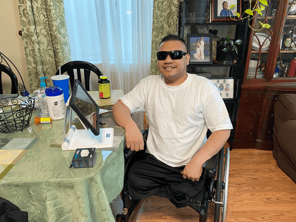 A man in a white t-shirt and sunglasses sits in a wheelchair at a table with various items in a living room. 